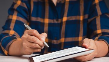 Geschäftsmann im karierten Hemd, der mit einem Stift ein digitales Dokument auf einem Tablet unterzeichnet und mit Anmerkungen versieht und eine detaillierte Vertragsanalyse zur Überprüfung und Genehmigung durchführt.