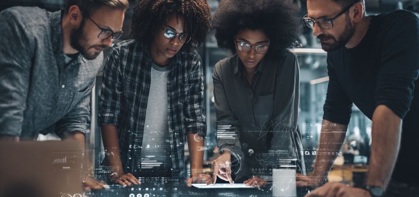 Un grupo diverso de colegas reunidos en torno a una mesa digital con pantalla táctil analizando juntos gráficos y documentos, utilizando un software de colaboración en contratos para agilizar las revisiones y aprobaciones de acuerdos.