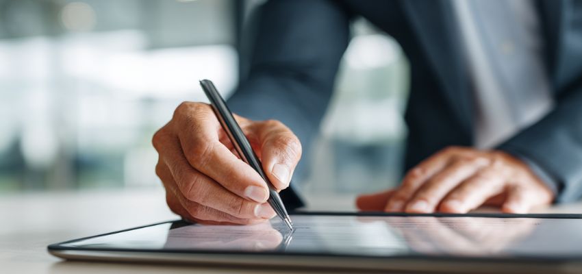 A professional in a suit uses a stylus to sign a contract addendum on a digital tablet.